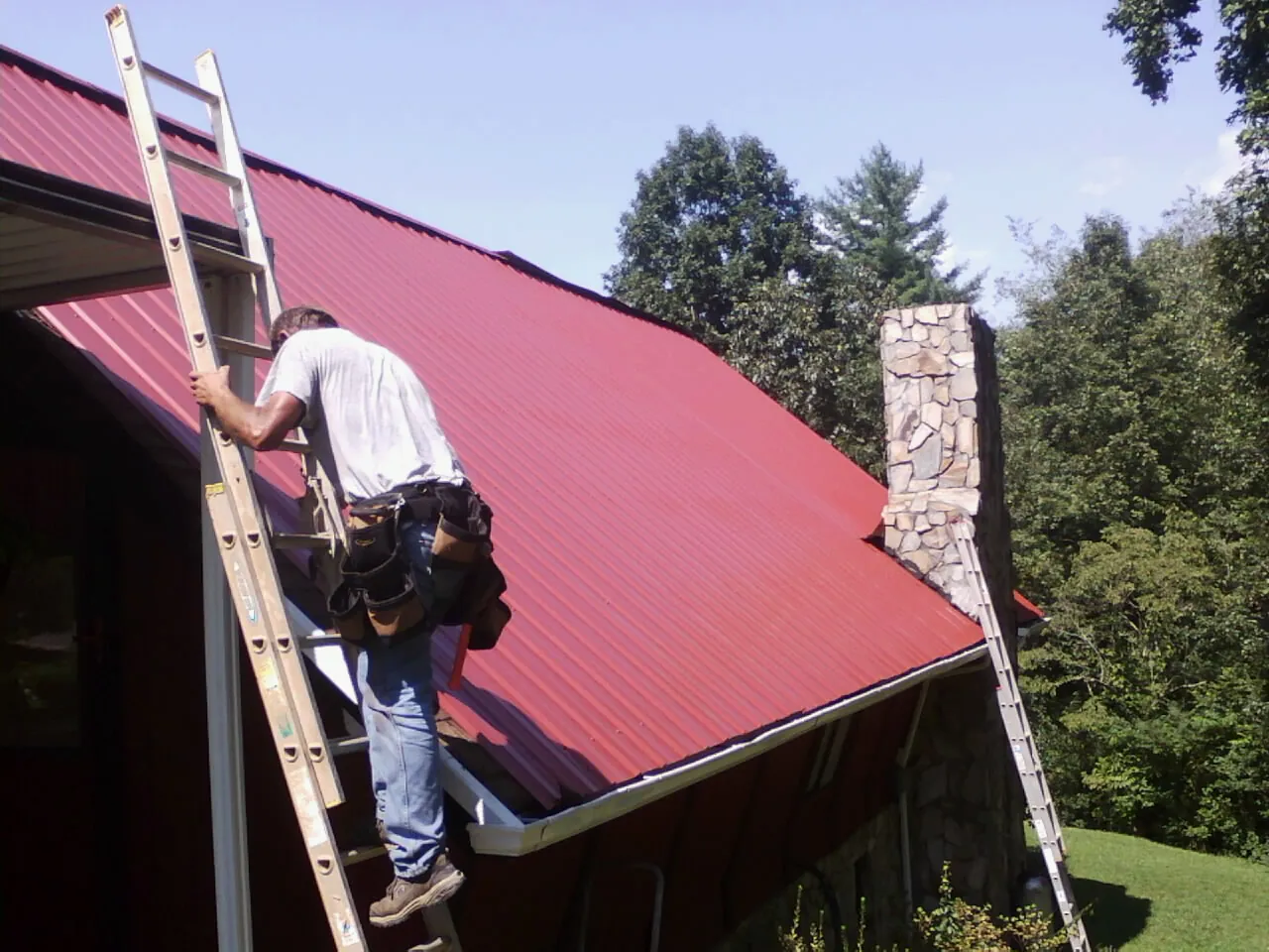 Roofer on a ladder beside a red metal roof and stone chimney — Soffit & Fascia Repair by Roofing Brothers in Viborg